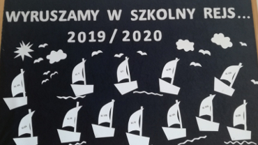 Tablica ogłoszeń z napisem "Wyruszamy w szkolny rejs 2019/2019" i białymi żaglówkami z papieru pływającymi poniżej